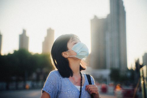 Free A woman wearing a face mask basks in sunlight amidst the cityscape of Hong Kong, capturing a moment of tranquility. Stock Photo
