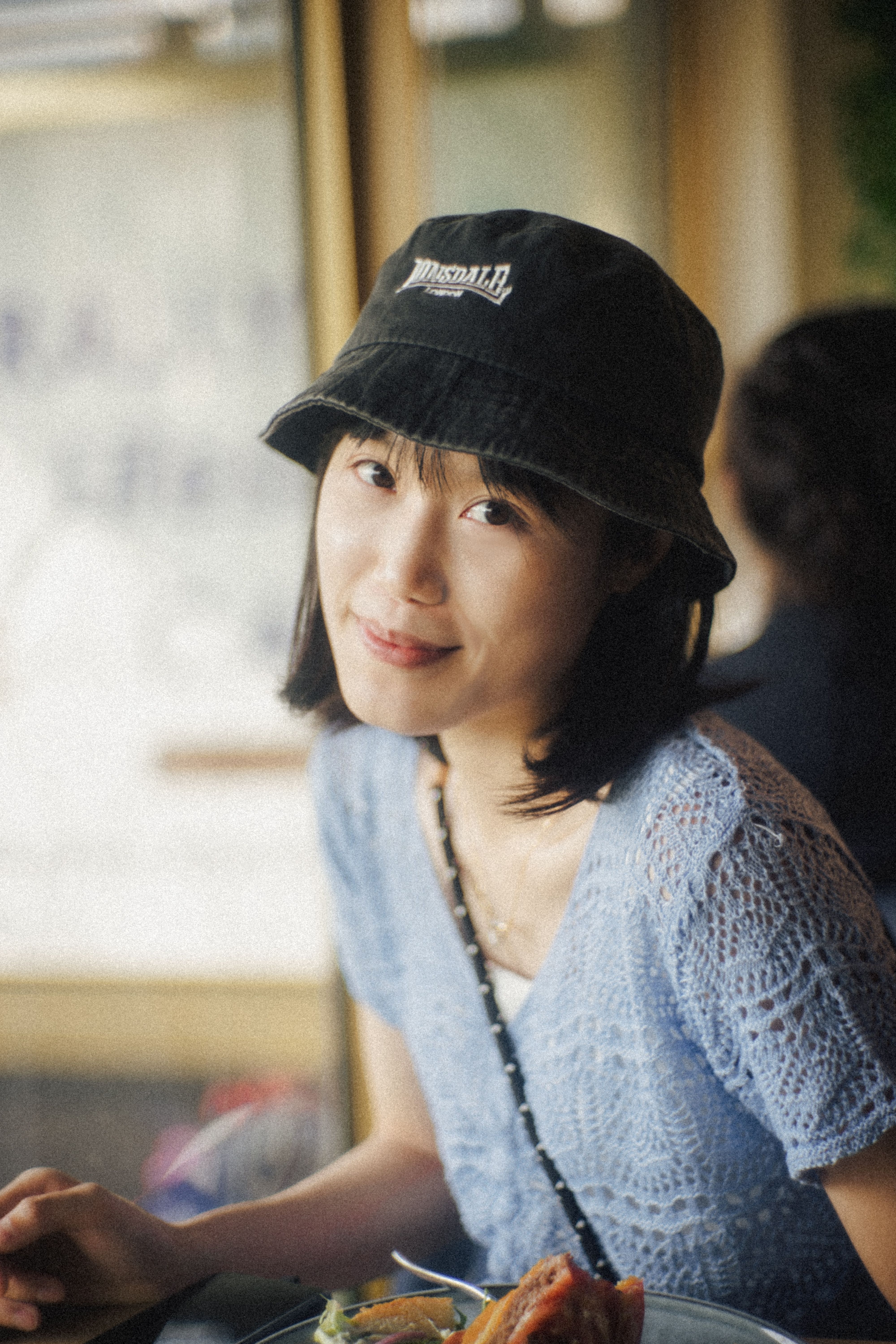 Smiling Asian woman wearing a bucket hat, sitting indoors with a relaxed vibe.