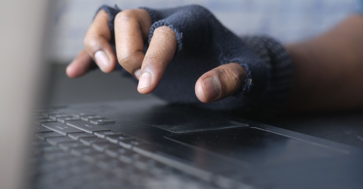 A person in gloves typing on a laptop keyboard, focusing on hands and keys in a dim-lit setting.