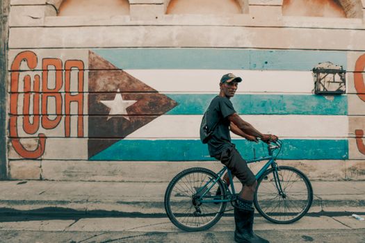 A man rides a bicycle by a vibrant Cuban flag mural in Havana.