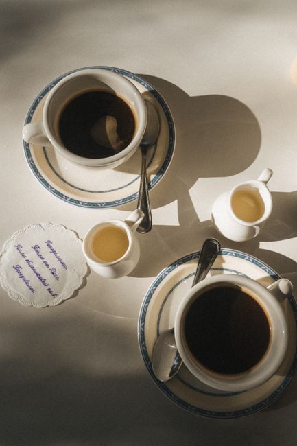 Overhead view of two coffee cups on saucers with milk jugs and a napkin in soft morning light.