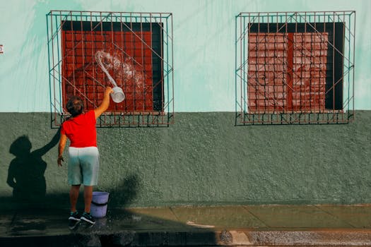 A woman in red shirt washing windows of a green house with a bucket and water.