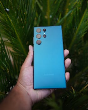 A hand holding a blue smartphone amidst green foliage, showcasing the device's sleek design in natural light.
