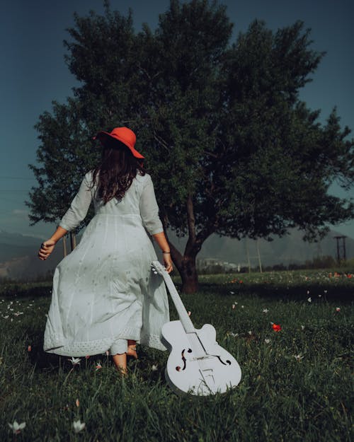 Free Back View of a Woman in White Dress Dragging a Guitar on Green Grass Field Stock Photo
