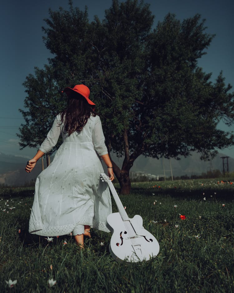 Back View Of A Woman In White Dress Dragging A Guitar On Green Grass Field