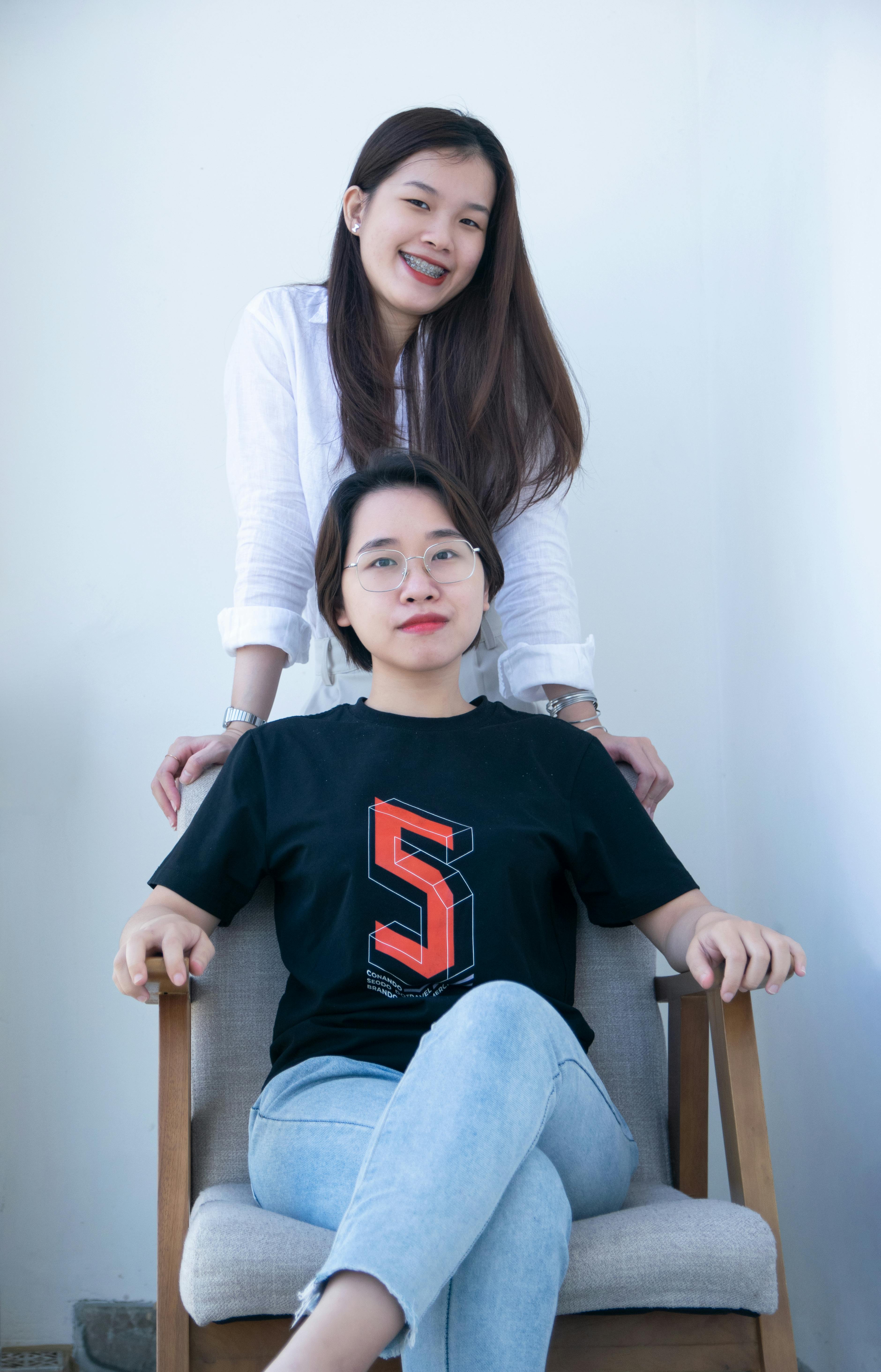 Two women, one sitting and one standing indoors, in casual clothing and smiling.