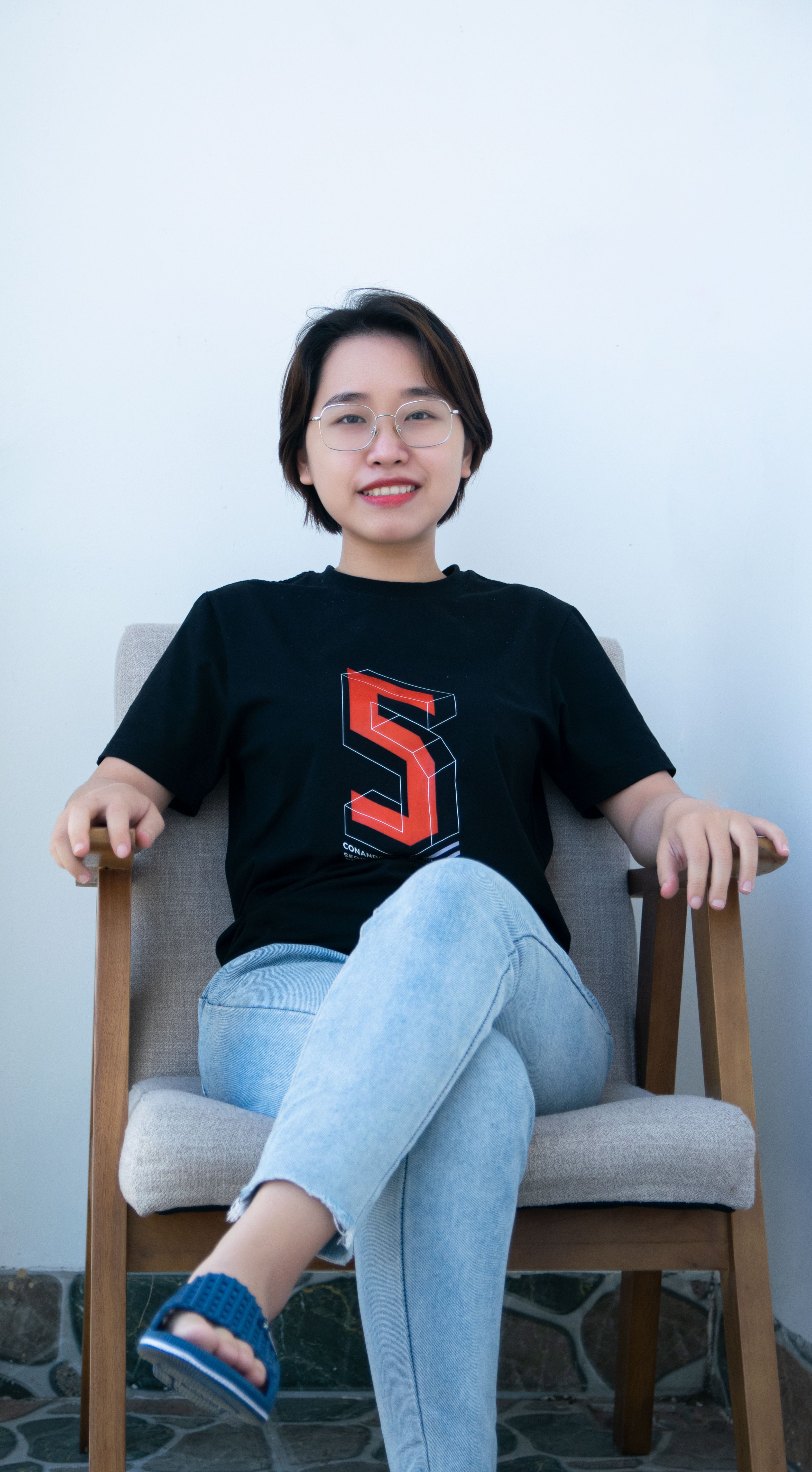 Asian woman in casual attire relaxing on a wooden chair indoors.