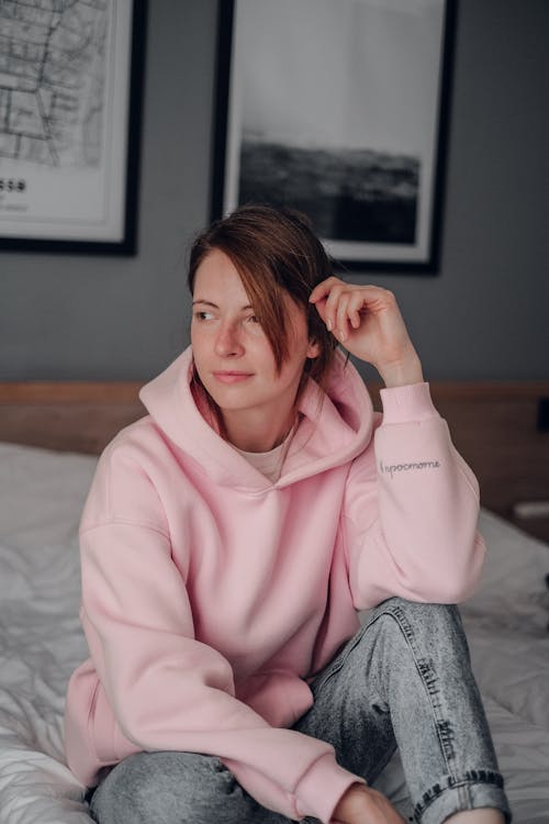 Free Woman in Pink Hoodie and Blue Denim Jeans Sitting on Bed Stock Photo