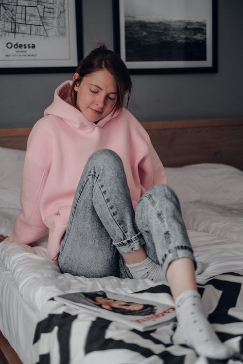 Free Woman in Pink Hoodie and Gray Pants Lying on Bed Stock Photo