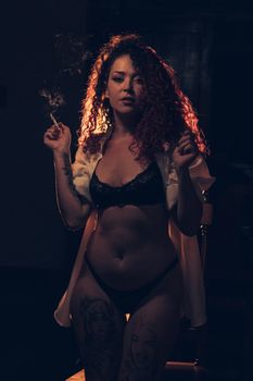 Artistic portrait of a woman in lingerie, standing and smoking in a dark setting, with tattoos visible.