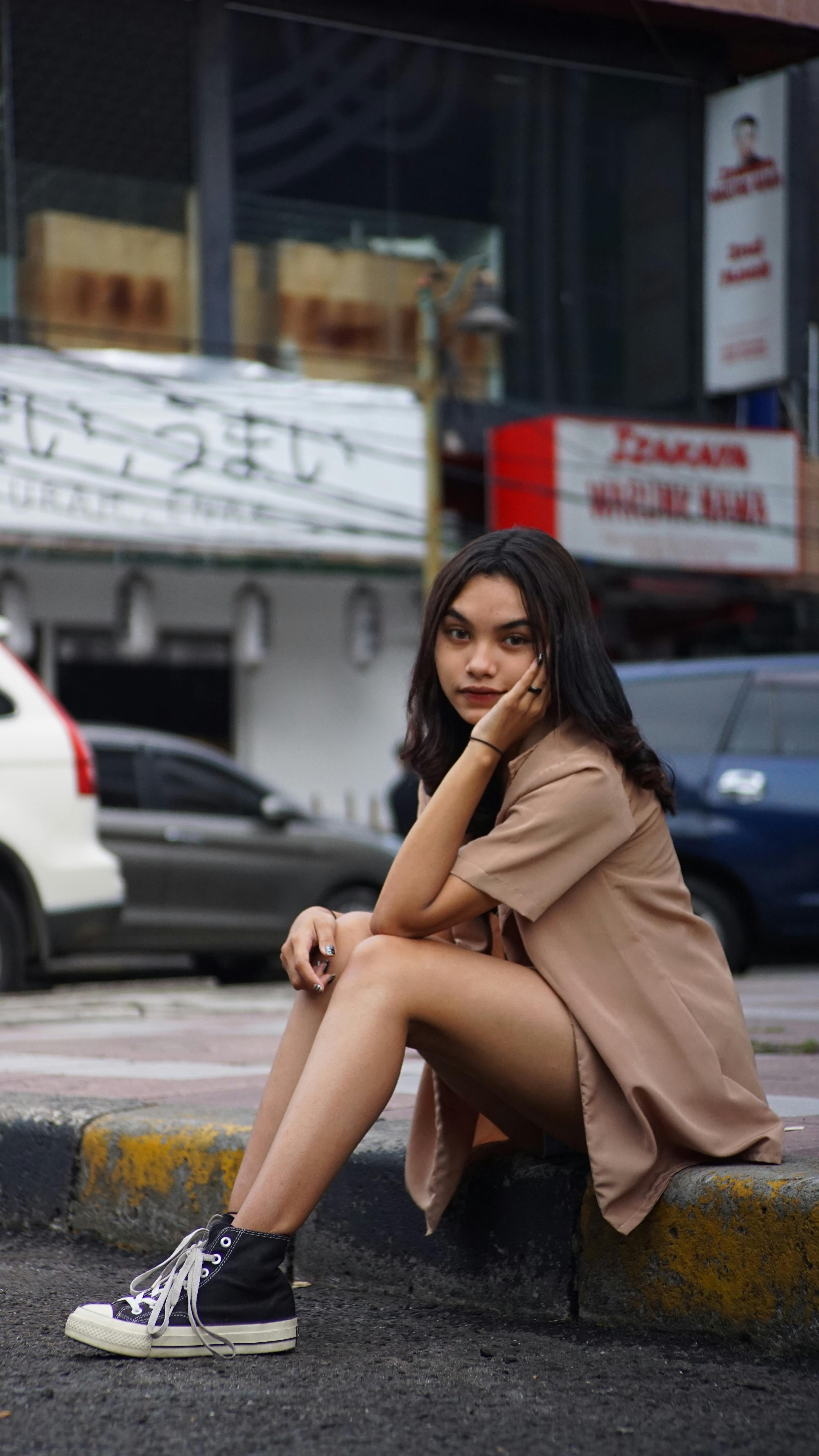 Stylish young woman in a brown shirt and Converse sneakers sitting on a city street curb, exuding a casual urban vibe.