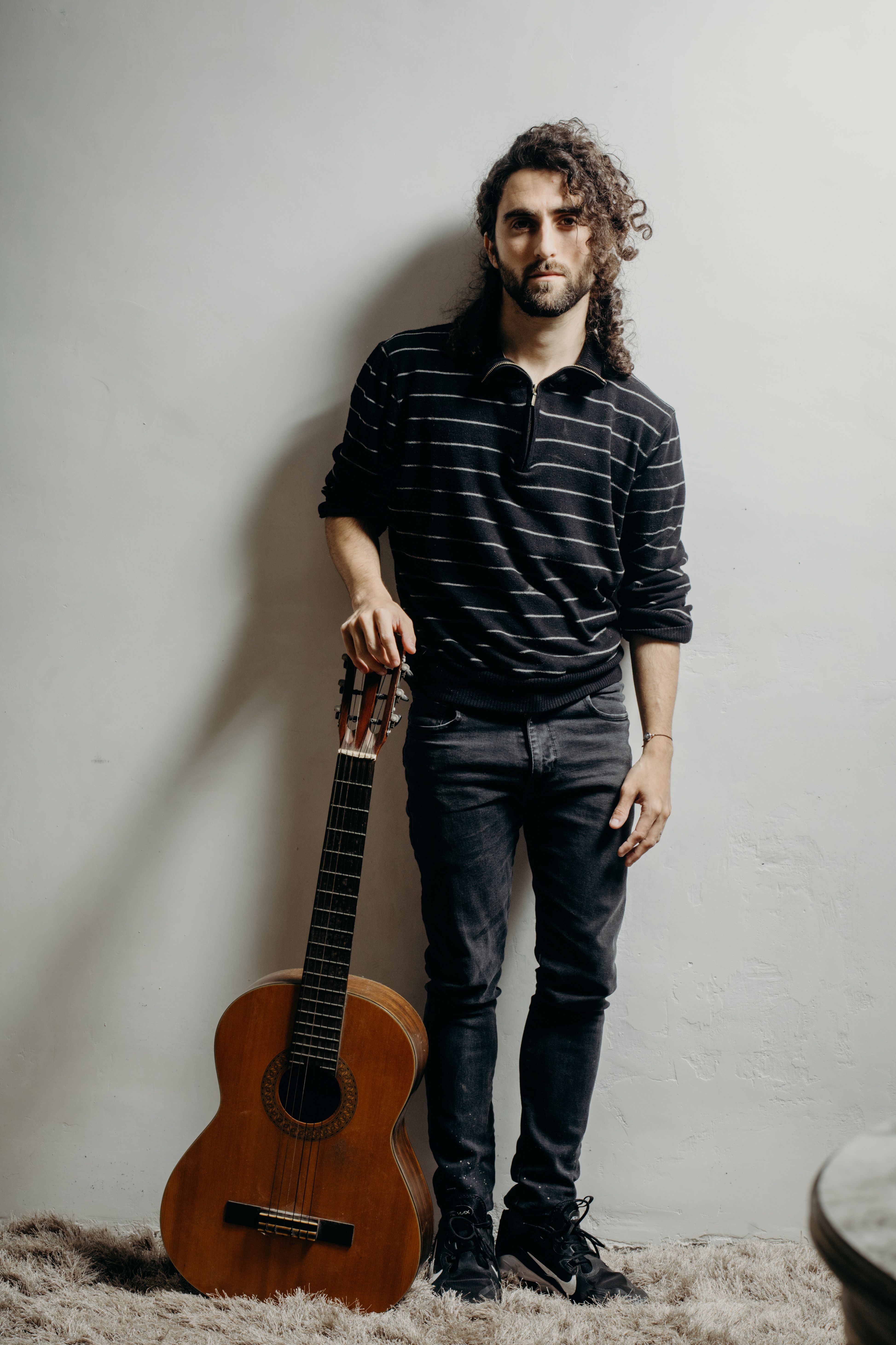 A man with curly hair and facial hair poses with a guitar indoors, showcasing casual fashion and musical talent.
