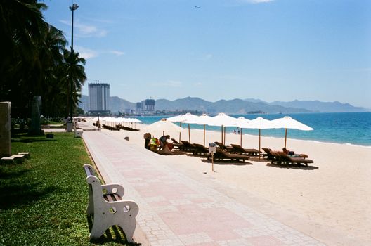Sunny day at a beach promenade with umbrellas and lounge chairs beside a turquoise sea.