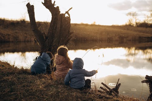 Children Sitting at a Lake Shore Playing with Grass