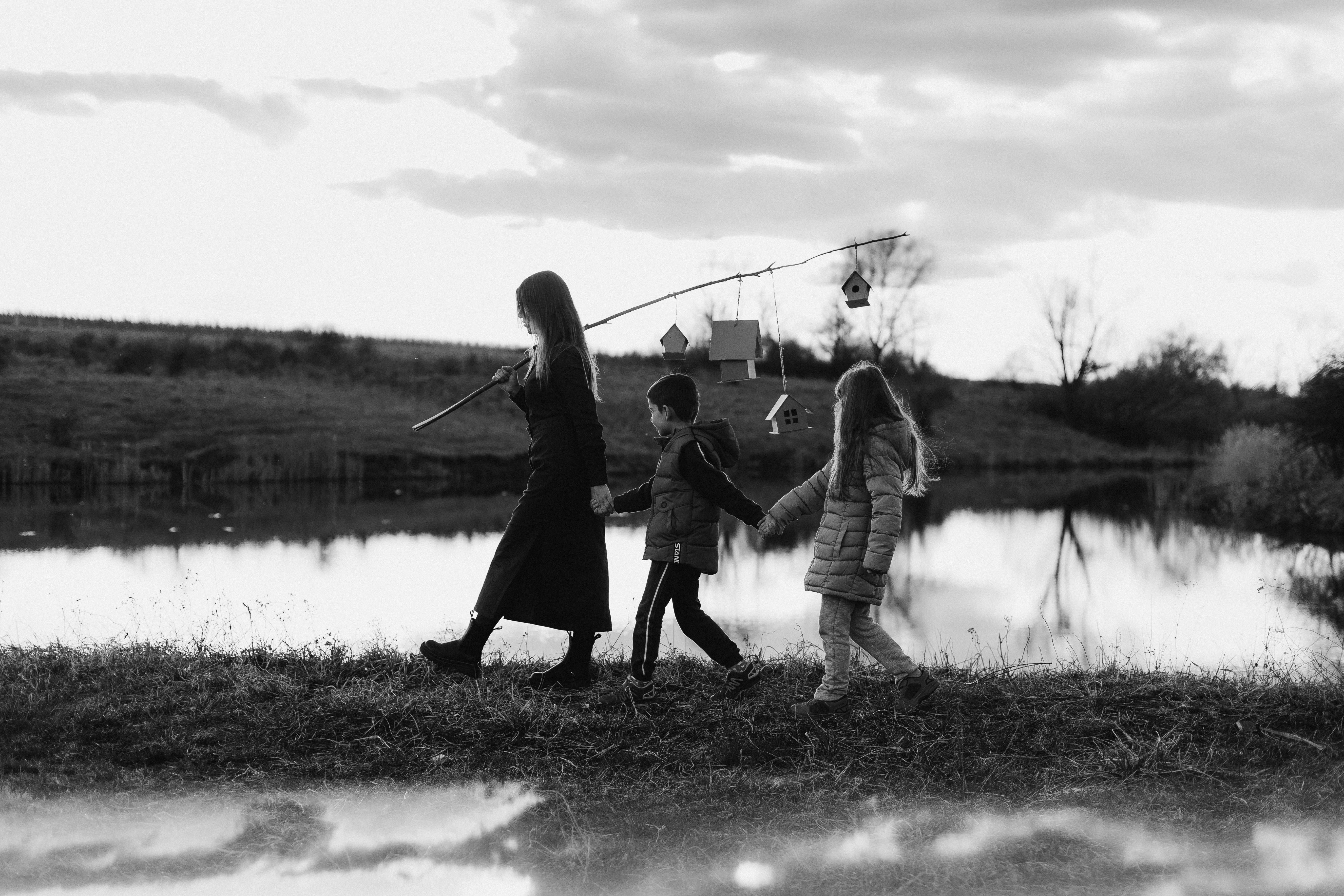 A mother and children walk by a tranquil lake, carrying birdhouses on a stick, in black and white.