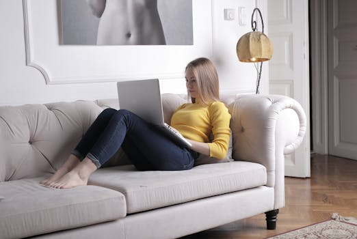 Young woman in yellow sweater using laptop on white sofa, creating a cozy and relaxed atmosphere.