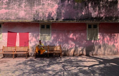 Free Elderly man reading newspaper on a wooden bench against a pastel building wall. Stock Photo