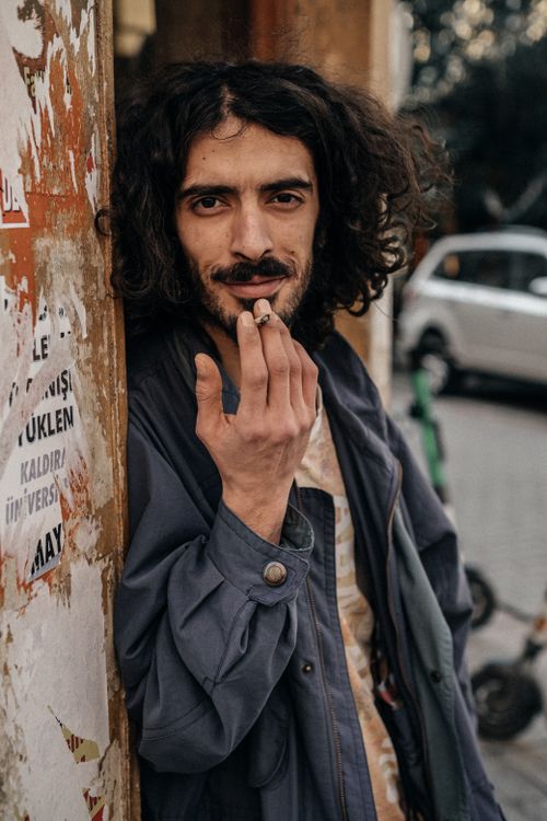 Free A man in Istanbul leans against a wall, wearing a blue jacket and smoking a cigarette. Stock Photo