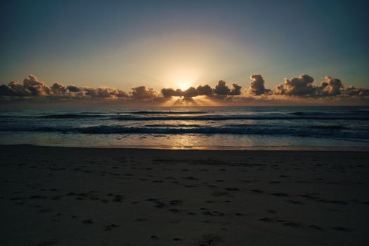 Stunning sunset view over a calm beach, with sun rays peeking through clouds on the horizon.