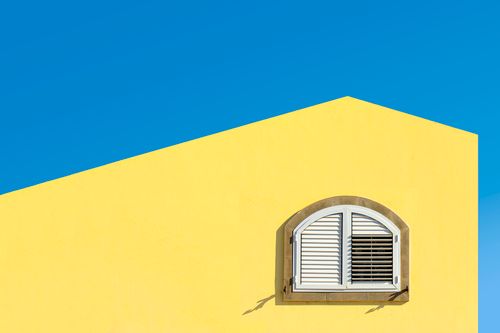 Free Bright yellow wall with an arched white shuttered window against a clear blue sky. Stock Photo