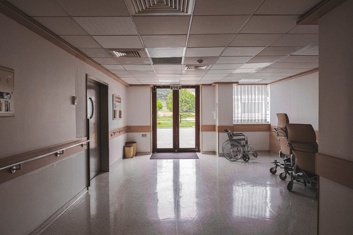 Free Hospital corridor with wheelchair, reflecting glass doors, and outdoor view. Ideal for healthcare themes. Stock Photo