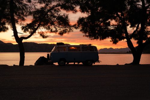 Free Silhouette of a vintage camper van parked beside a lake at sunset with dramatic skies. Stock Photo