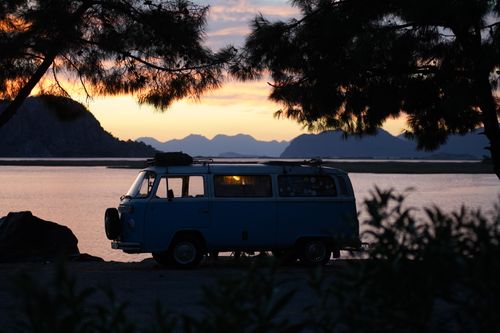 Free A vintage van parked by a calm lakeside, silhouetted against the sunset, surrounded by trees and mountains. Stock Photo