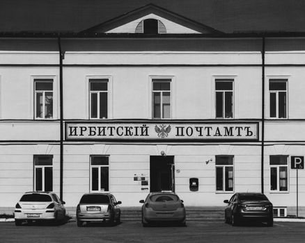 Grayscale facade of a historic building in Irbit, Russia, with parked cars.