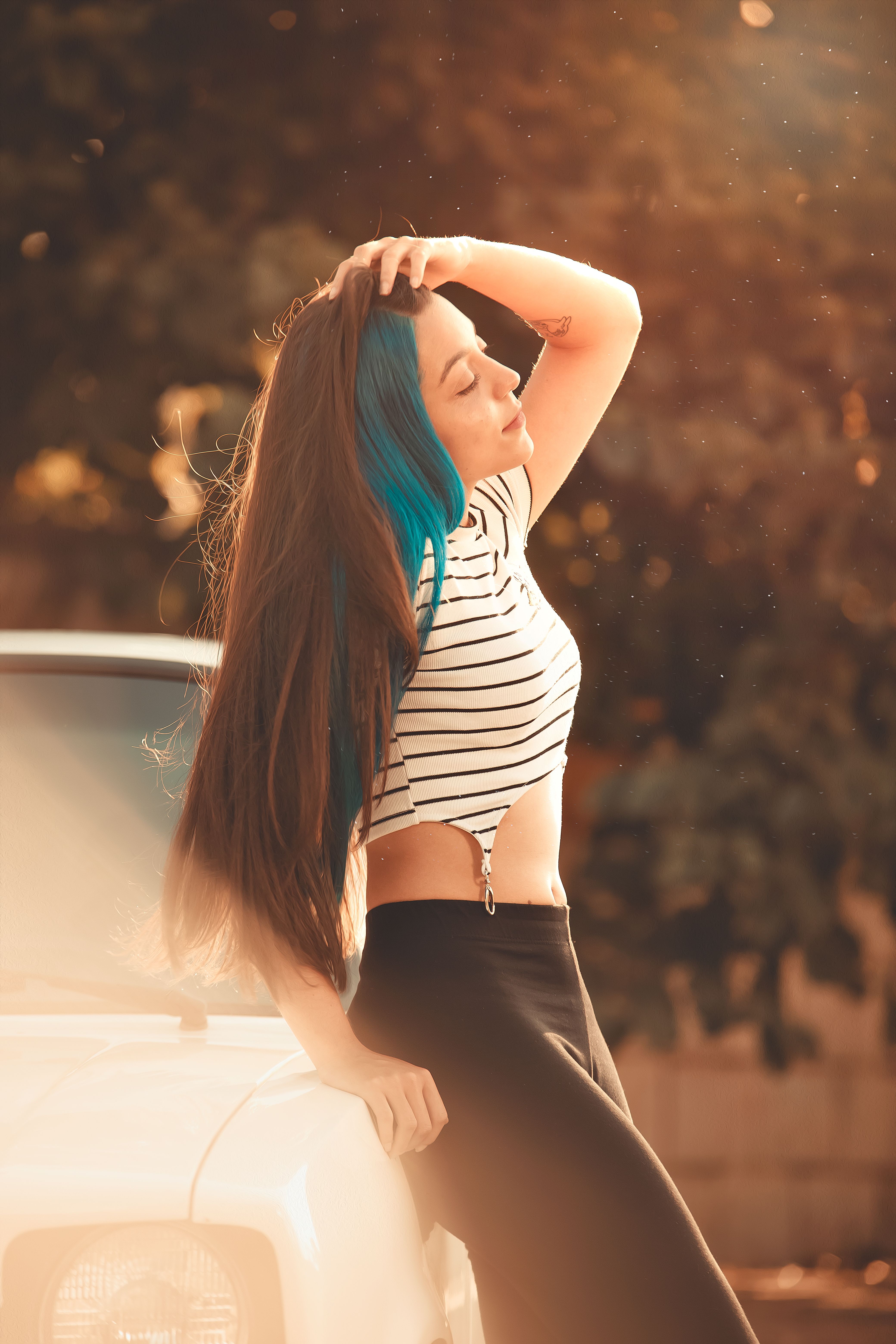 Outdoor portrait of a stylish young woman with blue highlights posing by a car in warm sunlight.