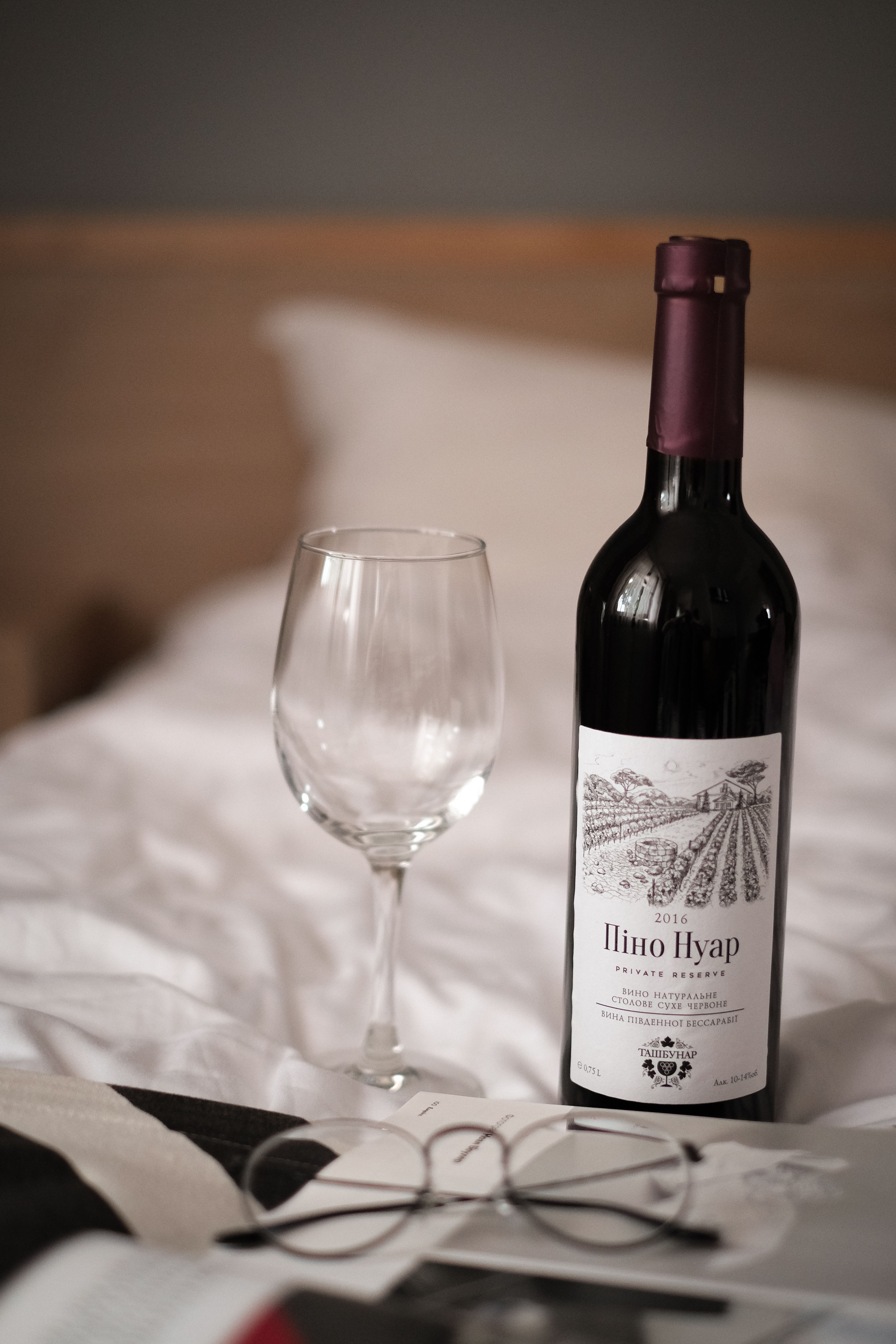A stylish still life of a wine bottle with a glass on a cozy bed setup.