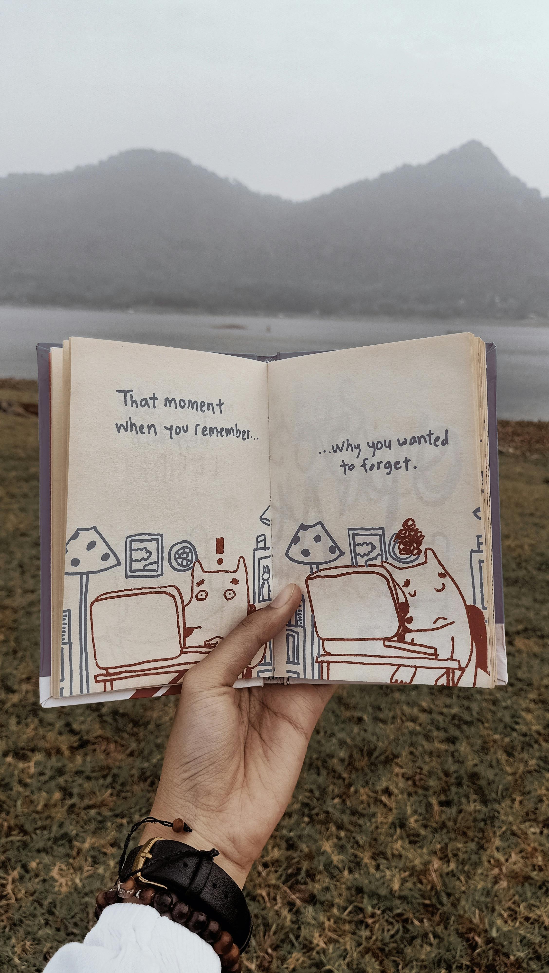 A person holds an open book showing an illustration in front of a scenic mountain and lake view.