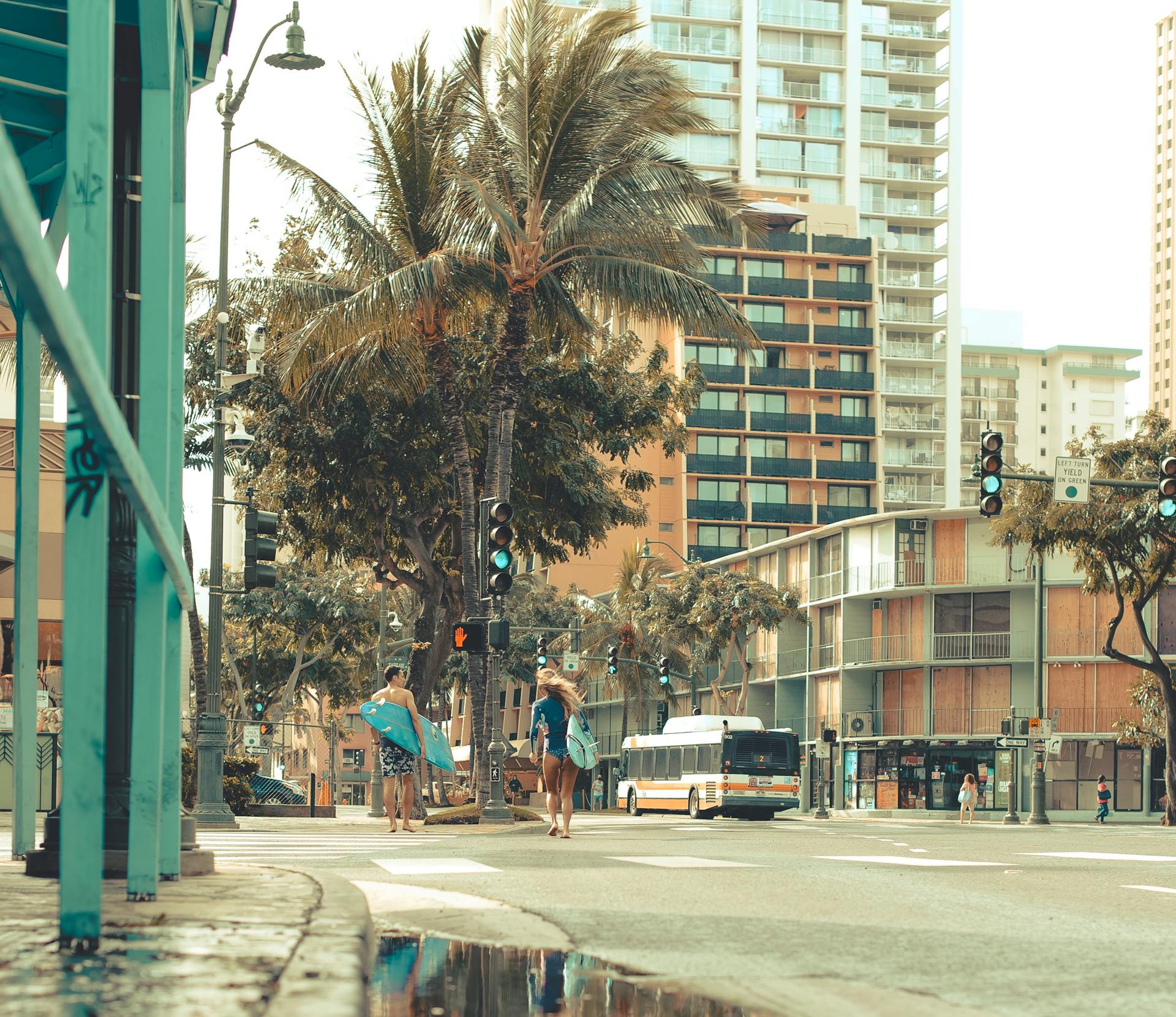 Surfers carrying boards cross a street surrounded by palm trees and city buildings in Honolulu.