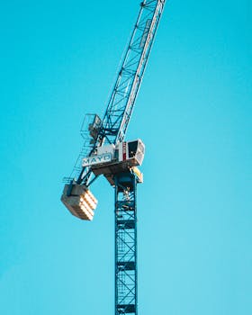 A vertical shot of a tower crane with 'MAYD' branding against a clear blue sky in Manchester, UK.