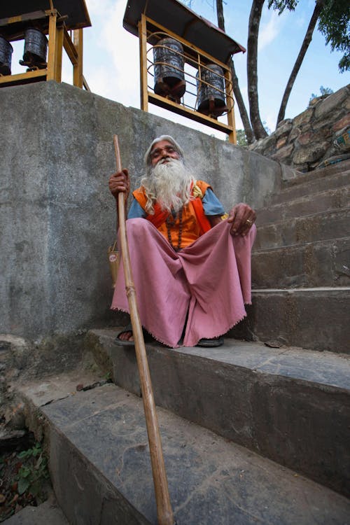 Senior Man Wearing Colourful Clothes Sitting with a Stick on Steps, and Bells in Background