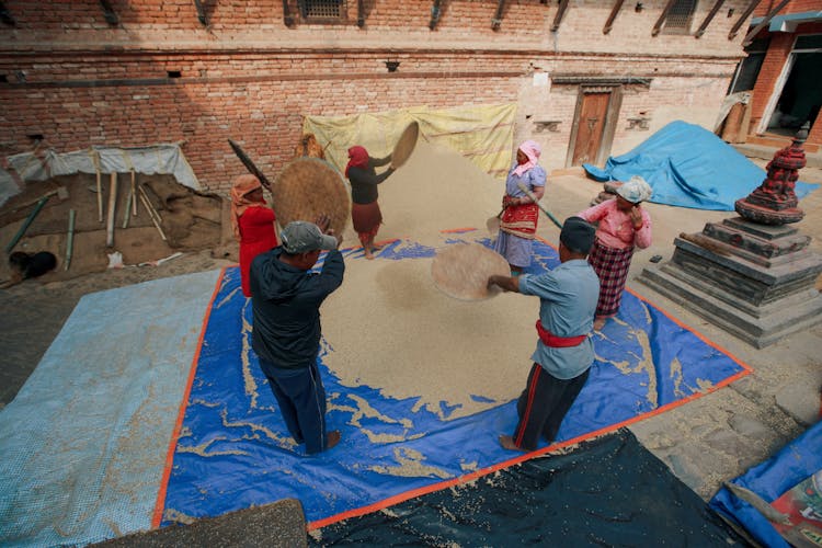 People Wearing Traditional Clothing Sifting Grain On A Yard