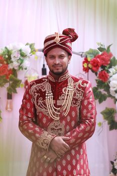 South Asian groom in ornate red and gold traditional attire, smiling amidst floral backdrop. Perfect for wedding themes.