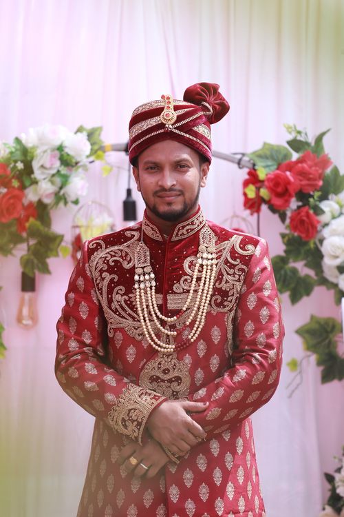 Free South Asian groom in ornate red and gold traditional attire, smiling amidst floral backdrop. Perfect for wedding themes. Stock Photo
