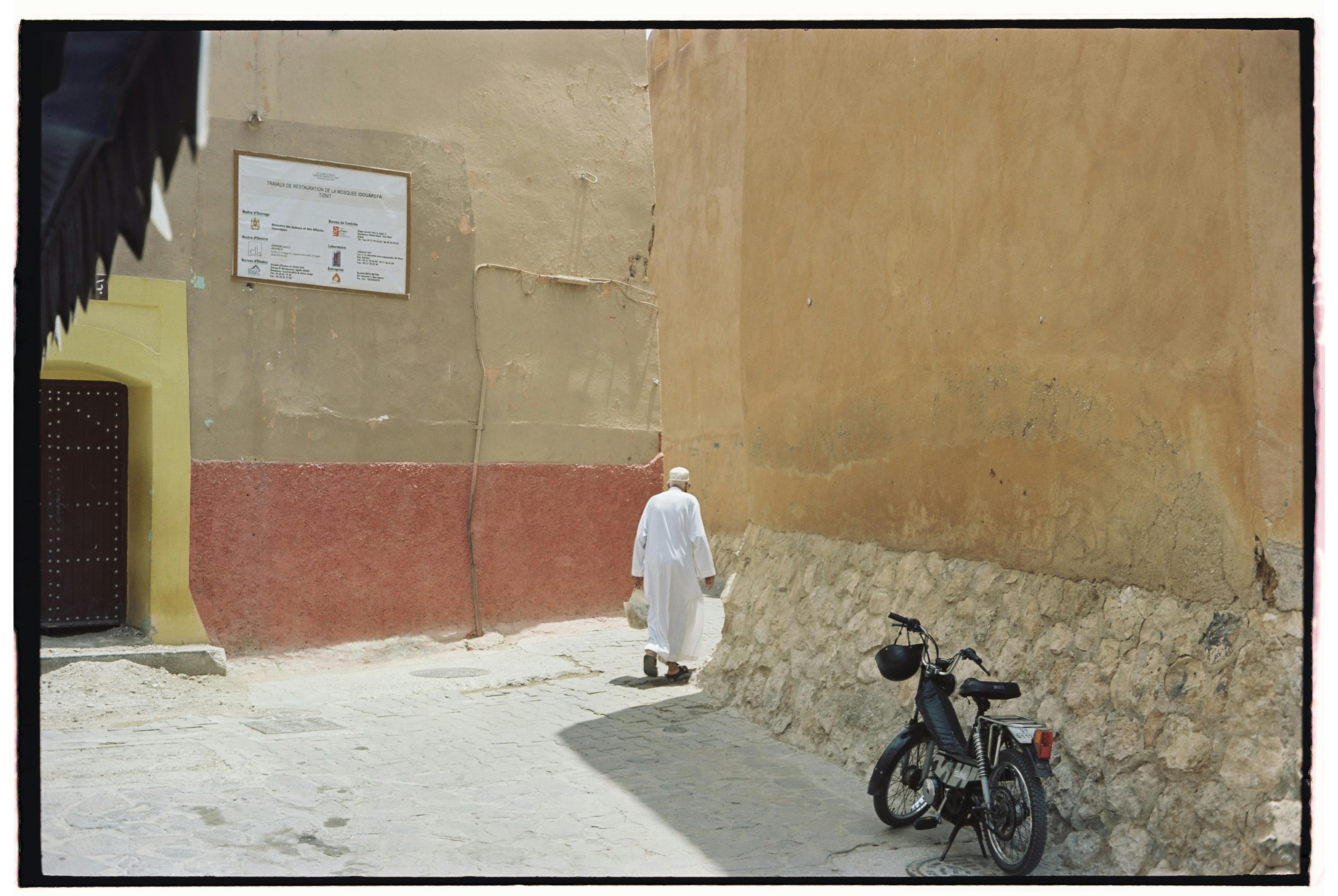 Man in a thobe walks down a narrow Moroccan alley with rustic walls and motorcycle.
