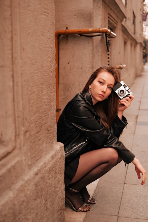 
A Woman in a Leather Jacket Holding a Camera while Squatting on a Sidewalk