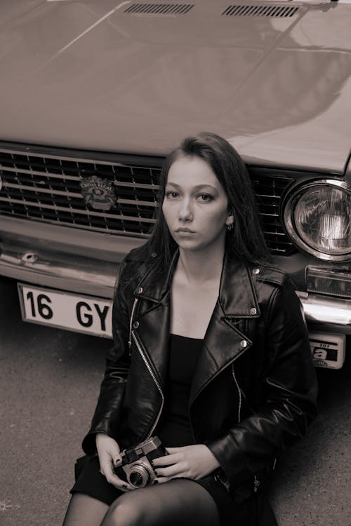 A Woman in Leather Jacket Holding a Camera while Sitting Beside a Car