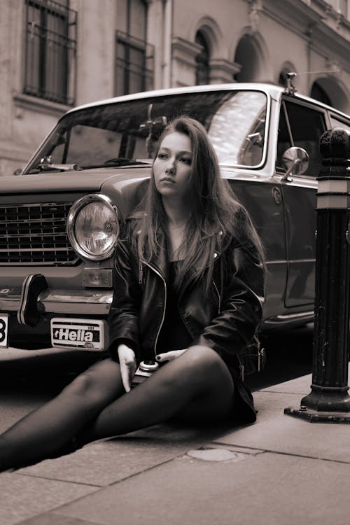
A Grayscale of a Woman in a Leather Jacket Sitting on a Curb