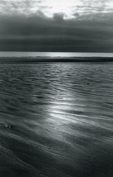Monochrome beach scene with rippled sand and calm ocean waves under cloudy sky.