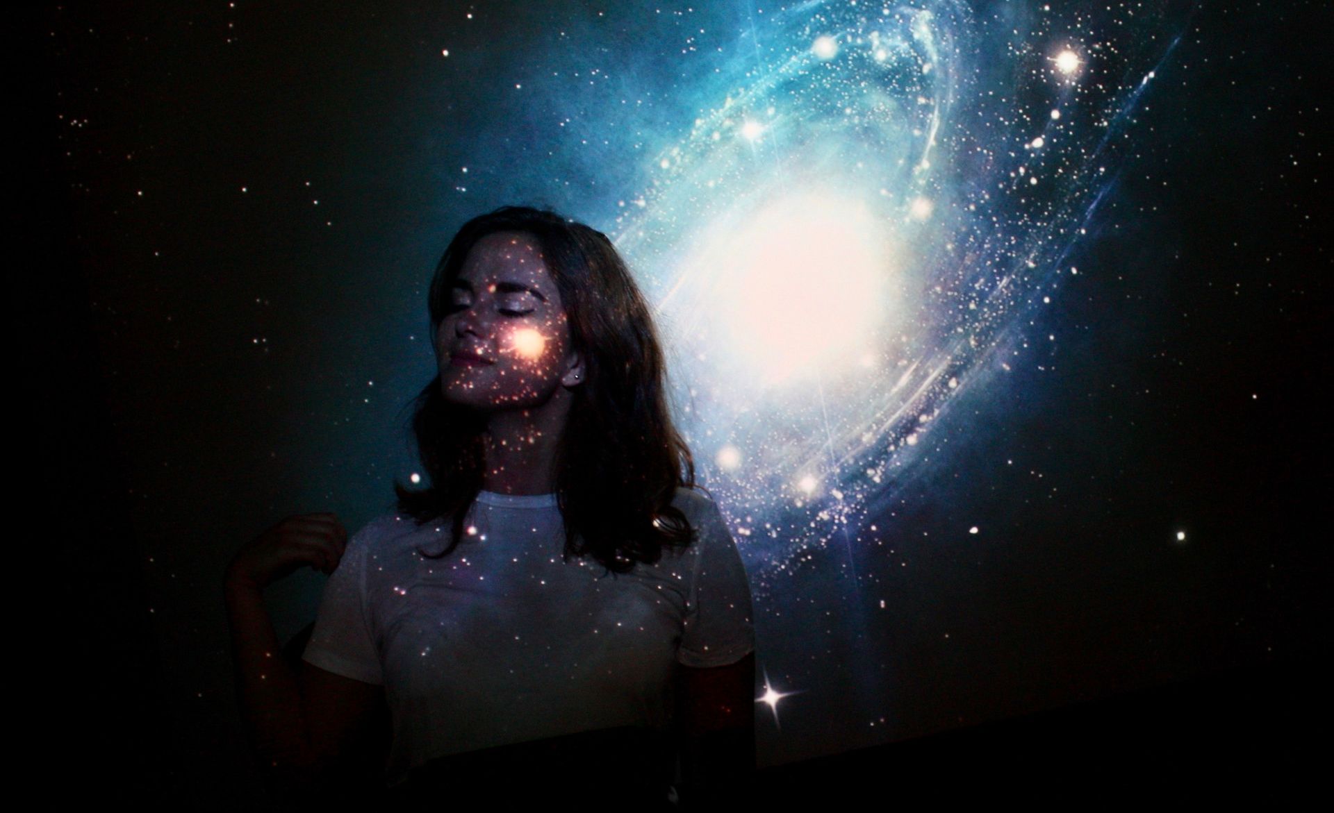 A young woman with eyes closed, standing against a galaxy projection, creating a serene and mystical atmosphere.