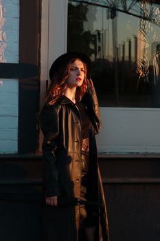 Fashionable woman in a black leather coat poses by a window during sunset.