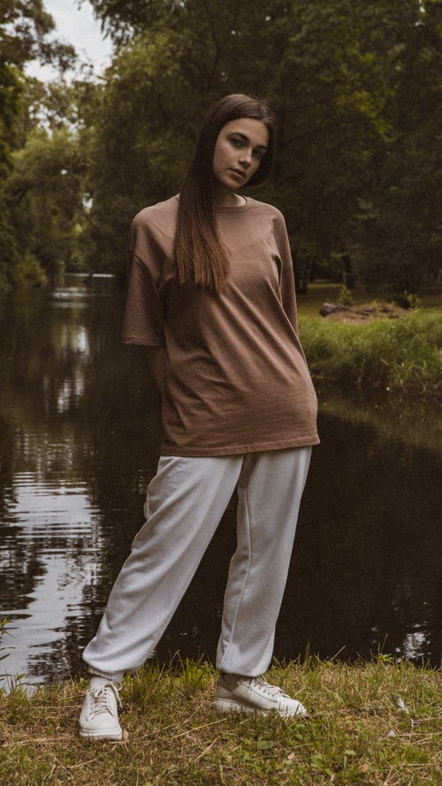 Free Teen girl in an oversized shirt standing by a stream in a tranquil park setting. Stock Photo