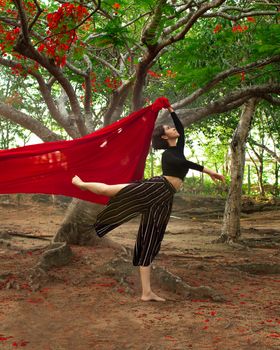 Woman dances gracefully among trees holding a red fabric, exuding artistic elegance.