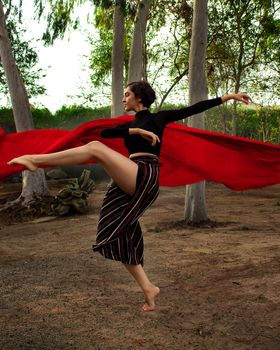 A woman performing a graceful dance outdoors with a vibrant red scarf among trees.