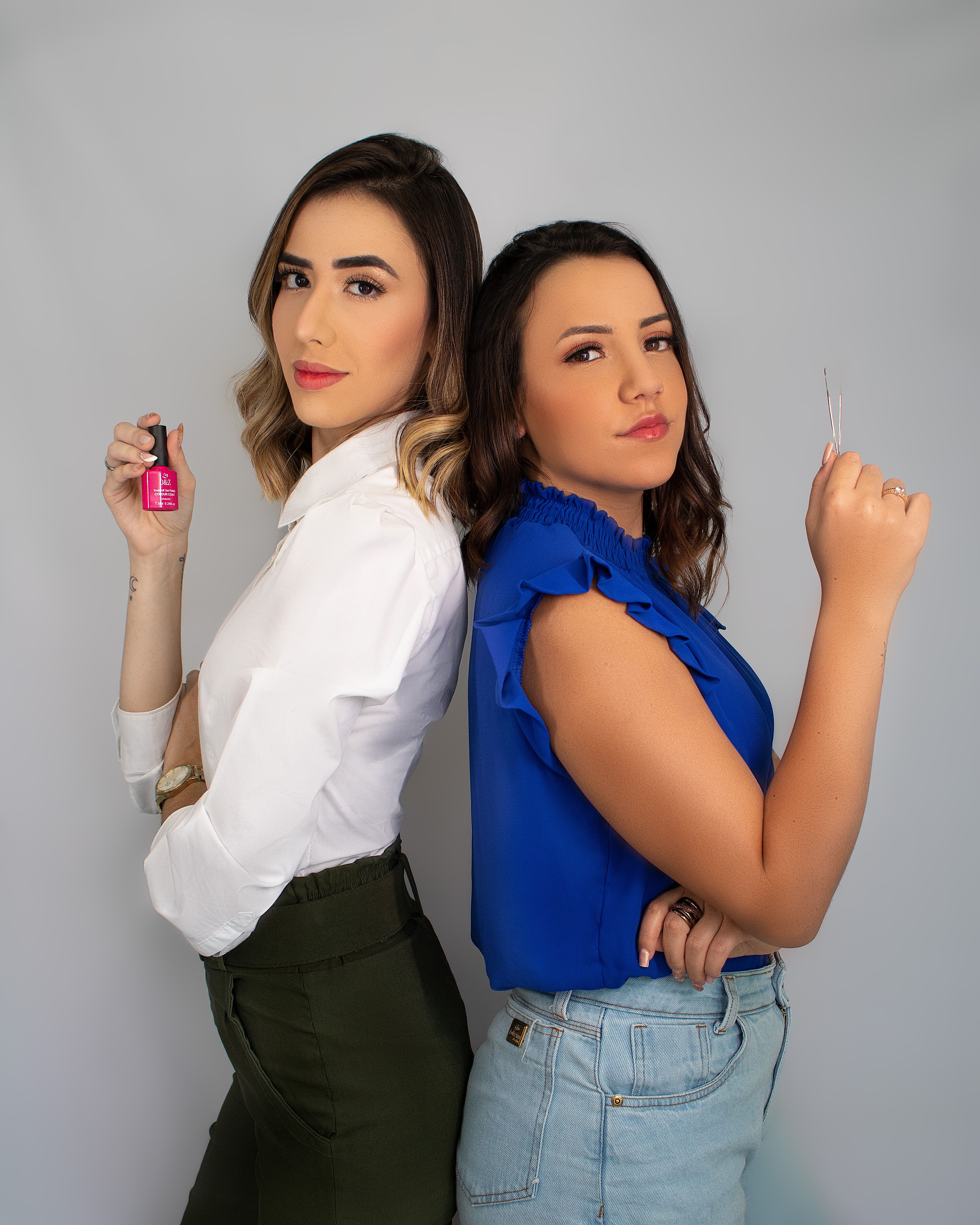 Two confident women pose back-to-back holding beauty tools in a studio setting.