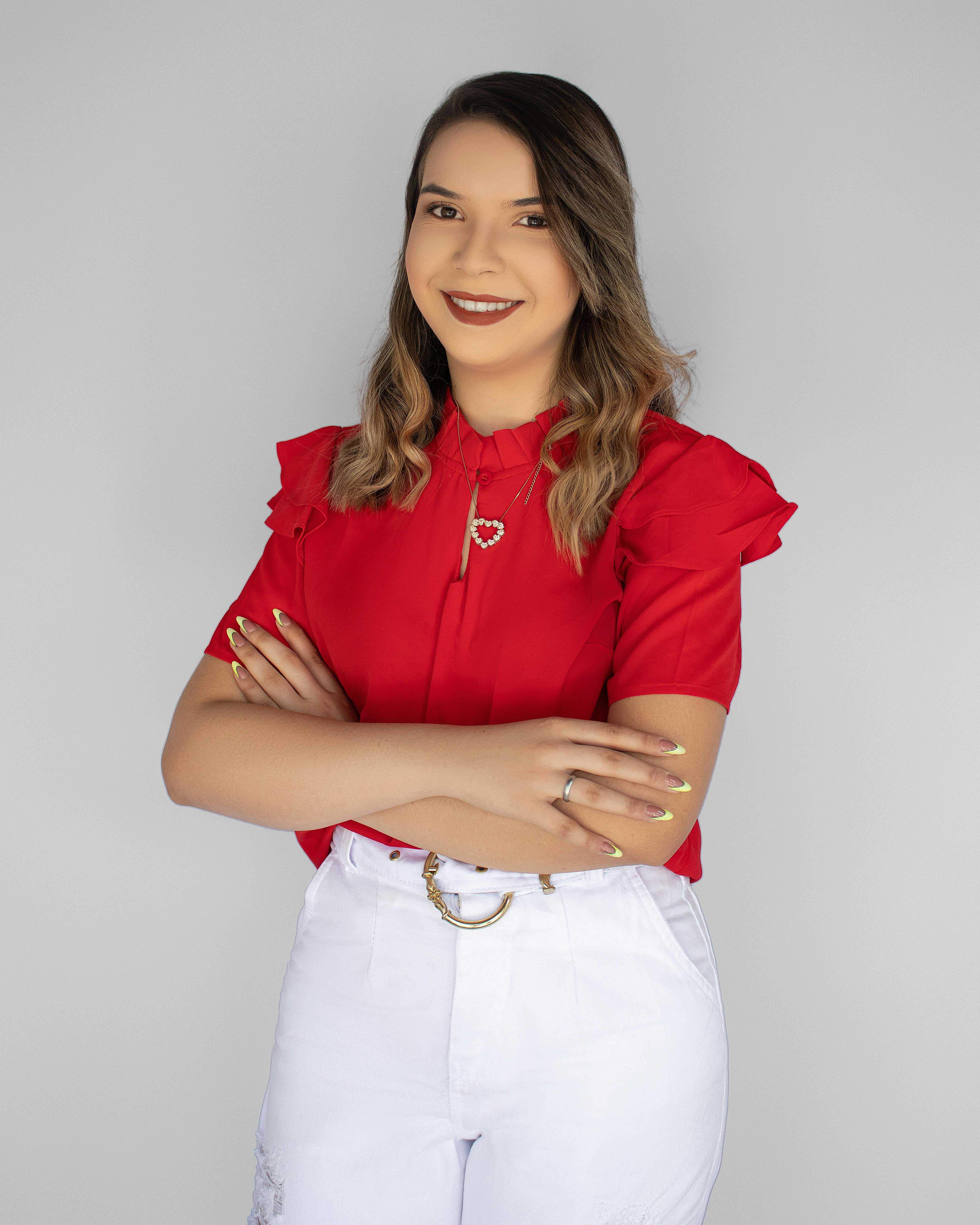 Portrait of a smiling woman in a red blouse and white pants, arms crossed, standing confidently in a studio setting.