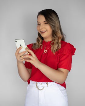 A woman in a red shirt smiles while using her smartphone in a studio setting, offering modern lifestyle vibes.